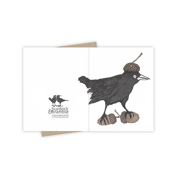Front and back view of the greeting card Grackle is foraging with a cap on top of his head and two acorns underfoot as he searches for food and provisions