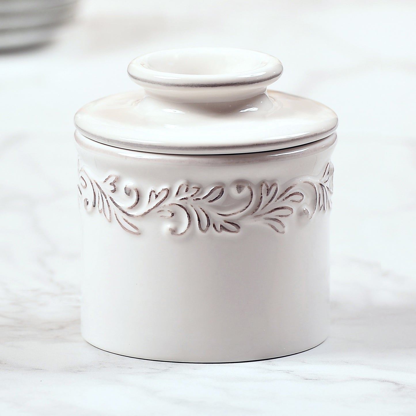 The Antique White linen Butter Bell Crock it has a design around the top of the base that is highlighted with a bronze glaze relief.