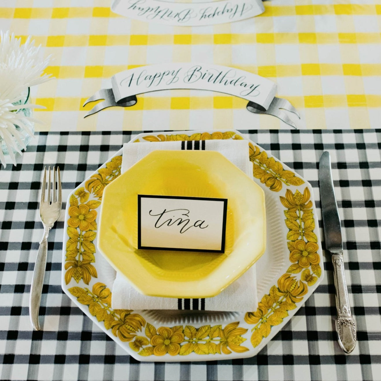 a photo of the black painted check paper placemat used in a table sitting. They have used yellow as the accent color in the dishes and table runner