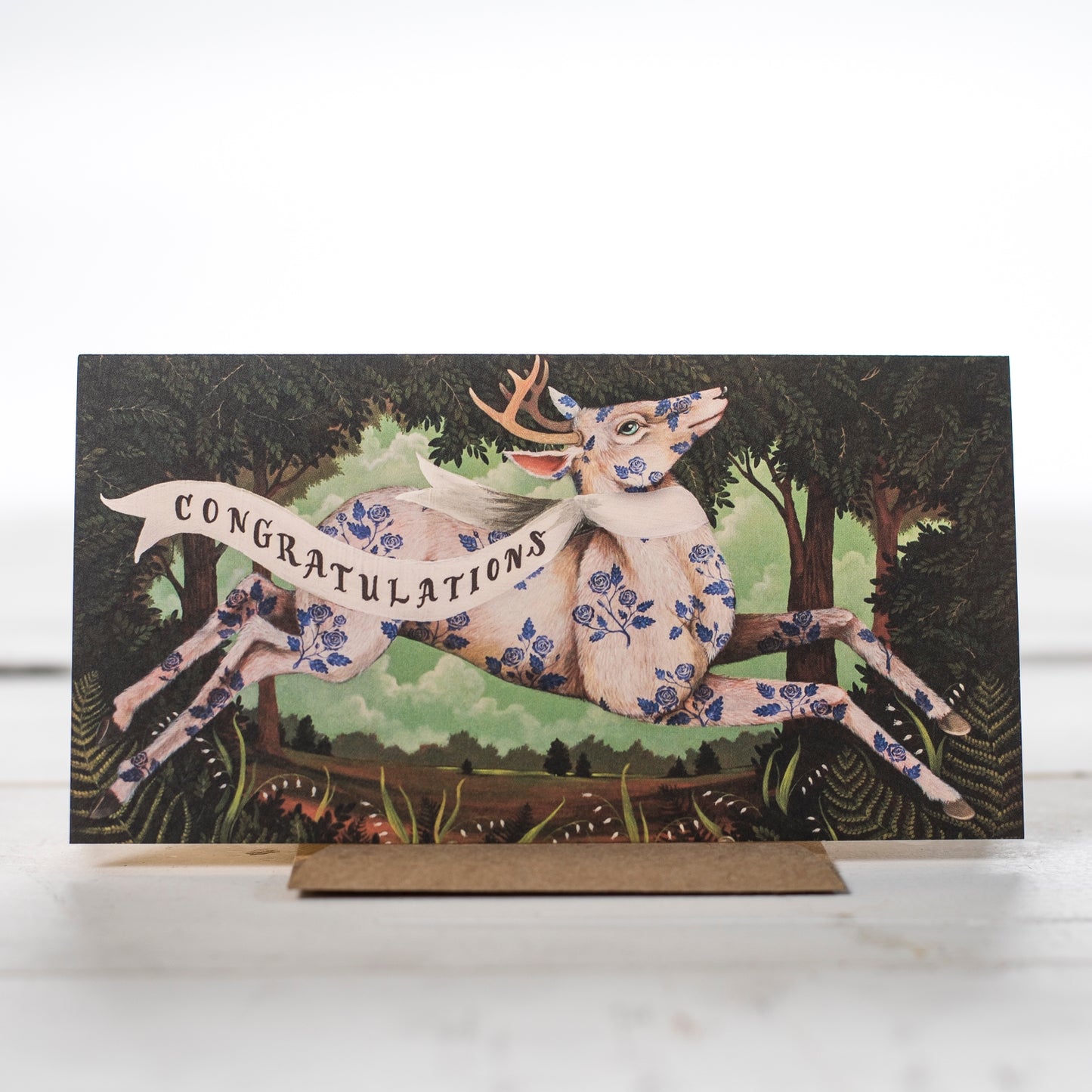 A deer leaping in a forrest covered with blue flowers printed on his fur wearing a banner that says Congratulations