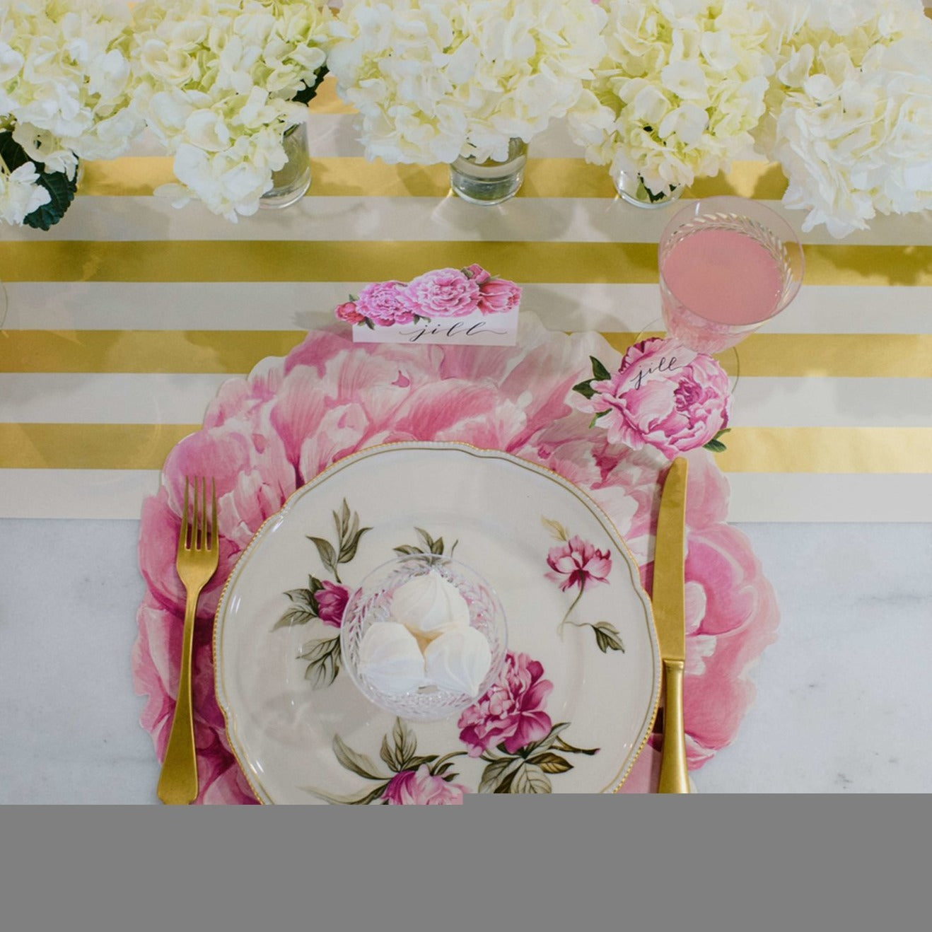 An image of the pink peony paper placemat used in a tablescape. Accented with gold silverware and white plates with pink flowers.