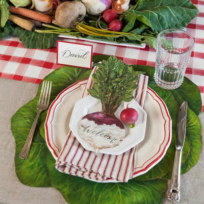 A picture of the green cabbage placemat being used at a table setting. It has white dishes trimmed with red  and a turnip place card.