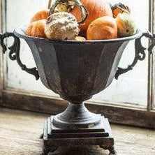 The distressed black metal urn with fall gourds arranged in it.