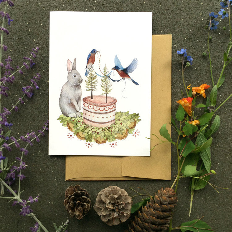 A rabbit setting with a birthday cake in front of him and two blue birds are decorating the cake with little trees and banners