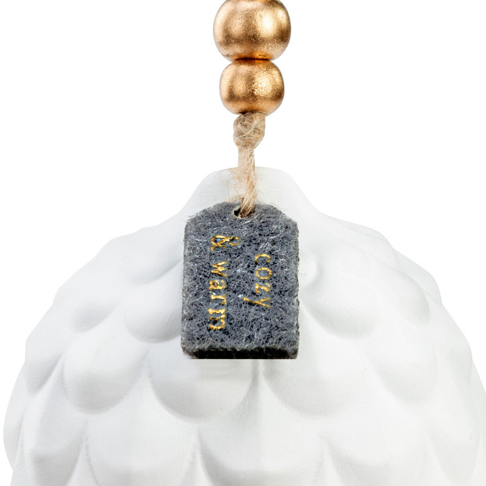 This is a close up picture of the top half of the Pine Cone Diffuser Ornament. It is a white ceramic pine cone dipped in gold at the tip, and is hung by a string with gold beads and a small sign saying "cozy and warm."