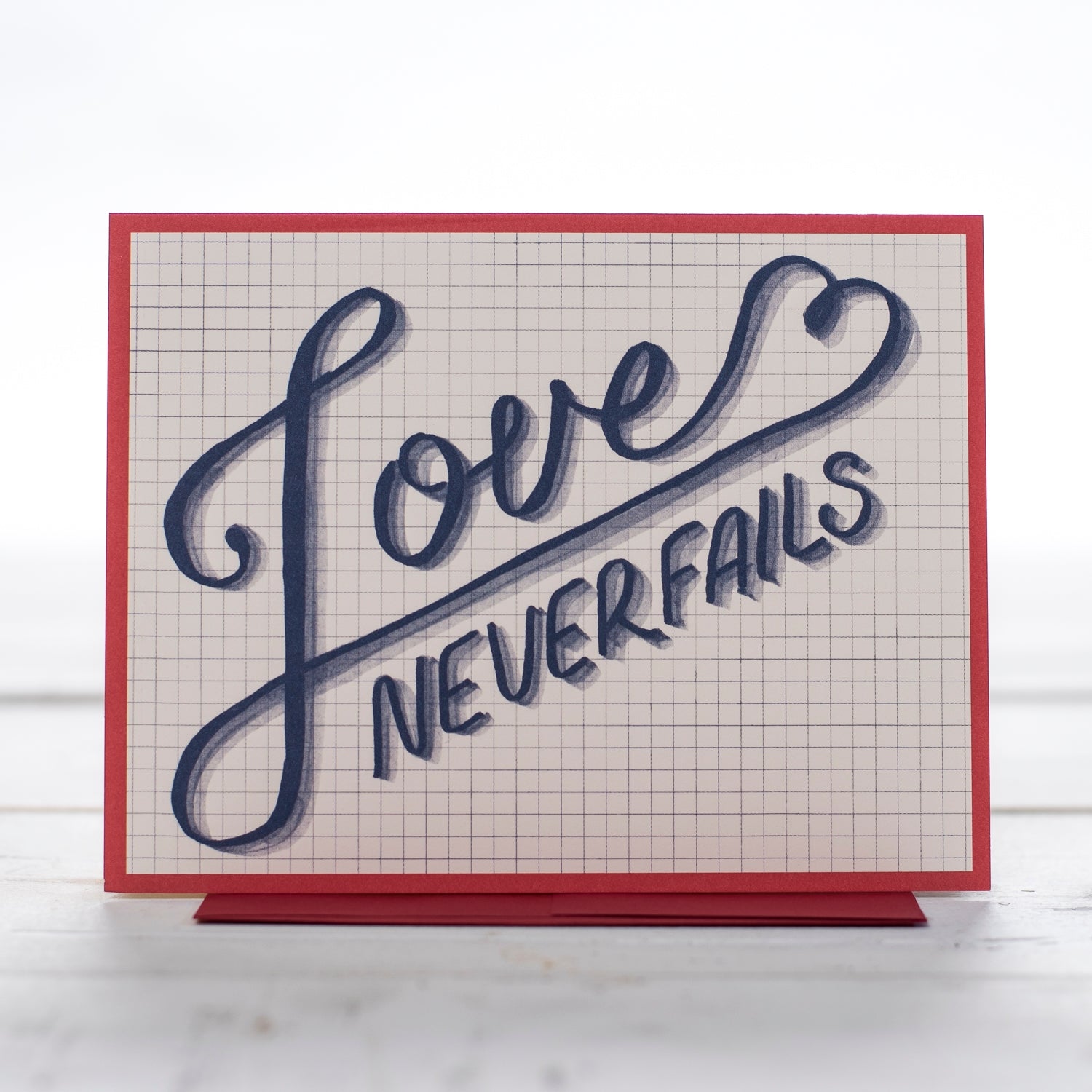 A greeting card with the words Love never fails in blue writing and a red boarder  on a grid back ground